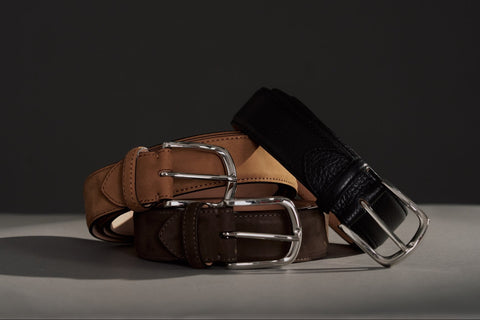 Three leather belts in brown, tan, and black, arranged in a stack with silver-tone metal buckles on a light gray surface against a dark gray background.